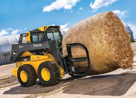 The words “ag material handling” are on top of a John Deere 332G Skid Steer with a bale hugger attachment moving a corn stalk round bale at a dairy farm.