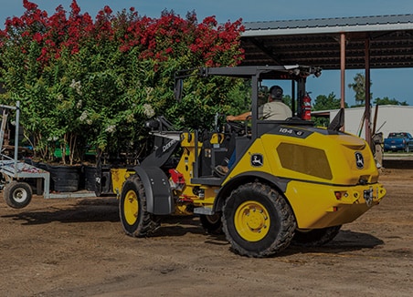 The word “landscaping” is on top of a John Deere 184 G-Tier Compact Loader moving a pallet full of Flowering Crape Myrtle in pots at a nursery.