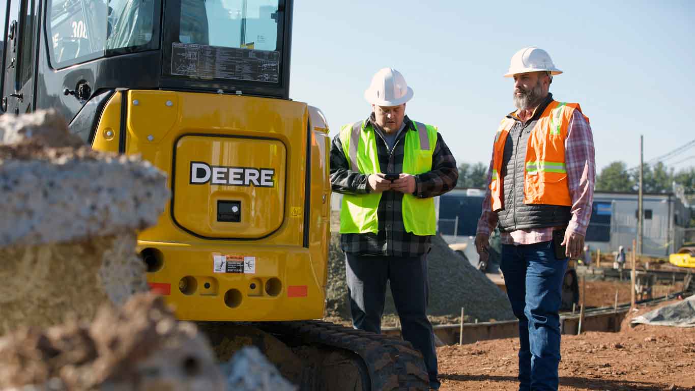 Two men in construction gear with one on their phone while standing next to John Deere construction equipment
