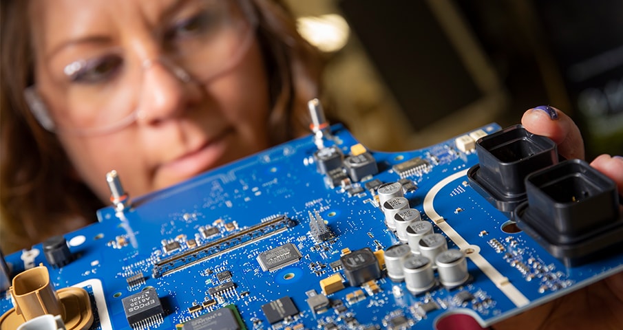 closeup of a blue circuit board with a person wearing safety goggles out of focus in the background