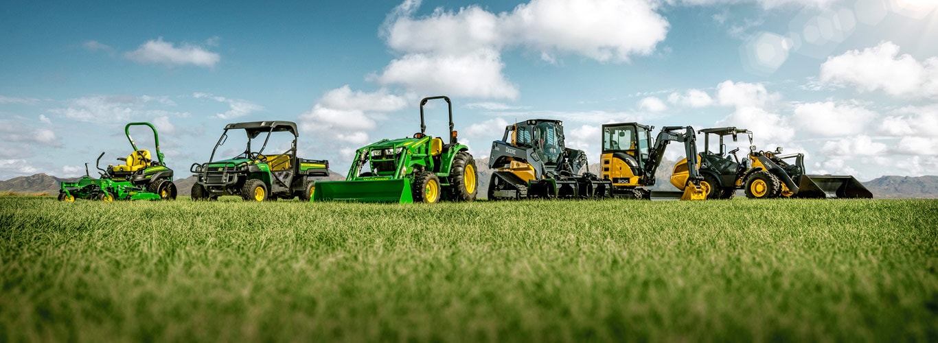 Lineup of John Deere landscaping and grounds care equipment in a grassy field with mountains in the distance