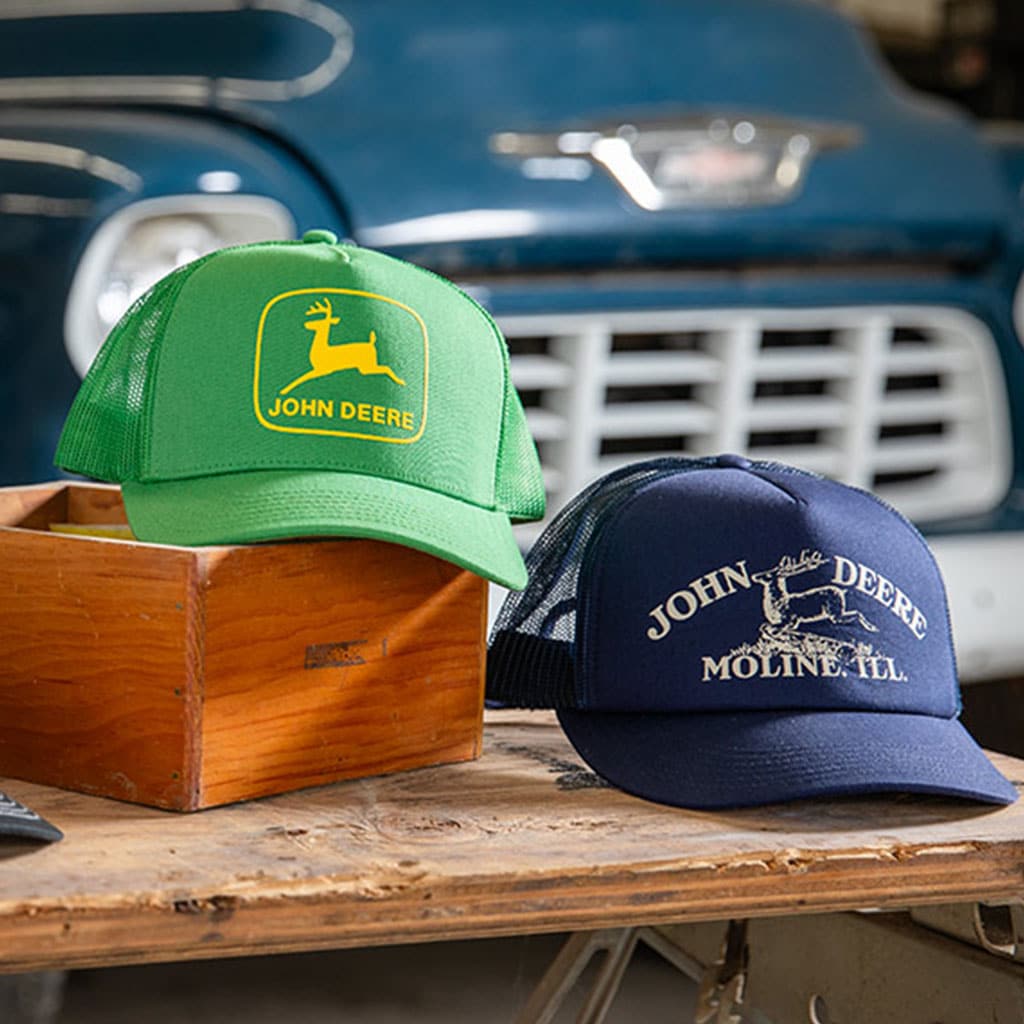 Two John&nbsp;Deere caps displayed on a wooden surface: a bright green mesh cap and a navy blue cap.