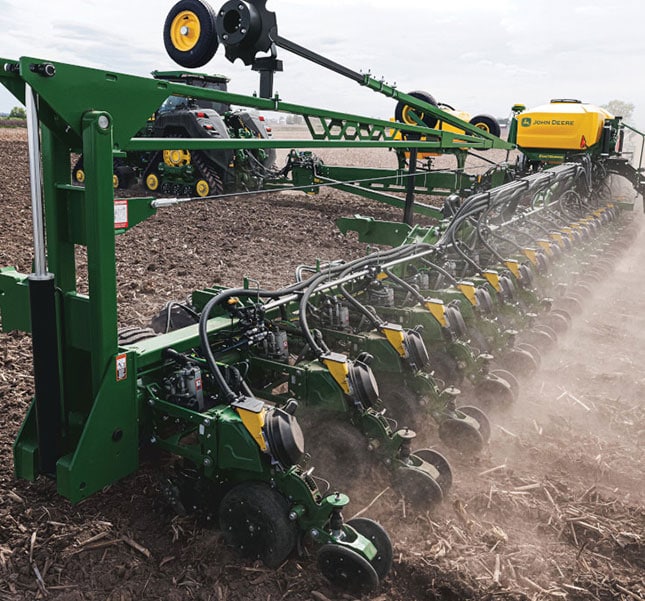 A green planter is working in a dusty field planting seeds in neat rows.