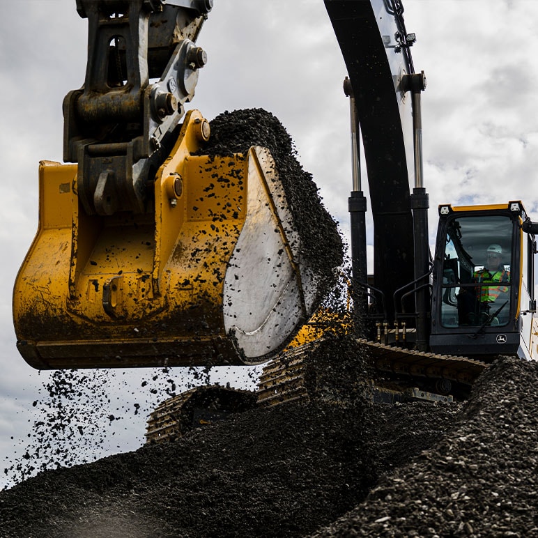 close up of an excavator bucket scooping dirt with the operator looking on in the background