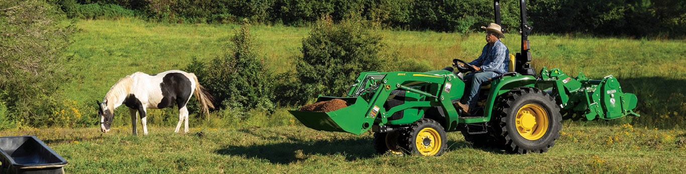 Person driving a 3 Series Compact Tractor across grass with dirt in the front-loader. A horse grazes nearby.