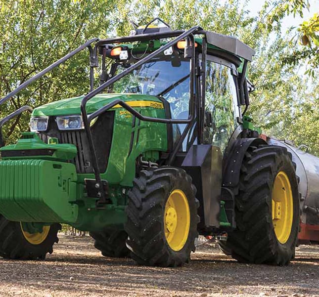 A green tractor pulling a sprayer through an orchard.
