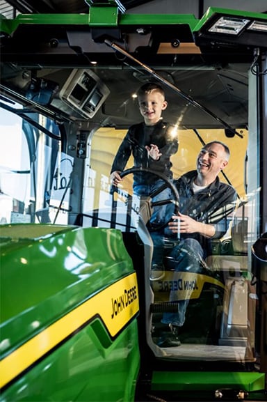 A father and his son sit inside a John Deere tractor cab smiling together