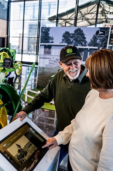 Two people experience John Deere history facts on a screen in front of a classic tractor