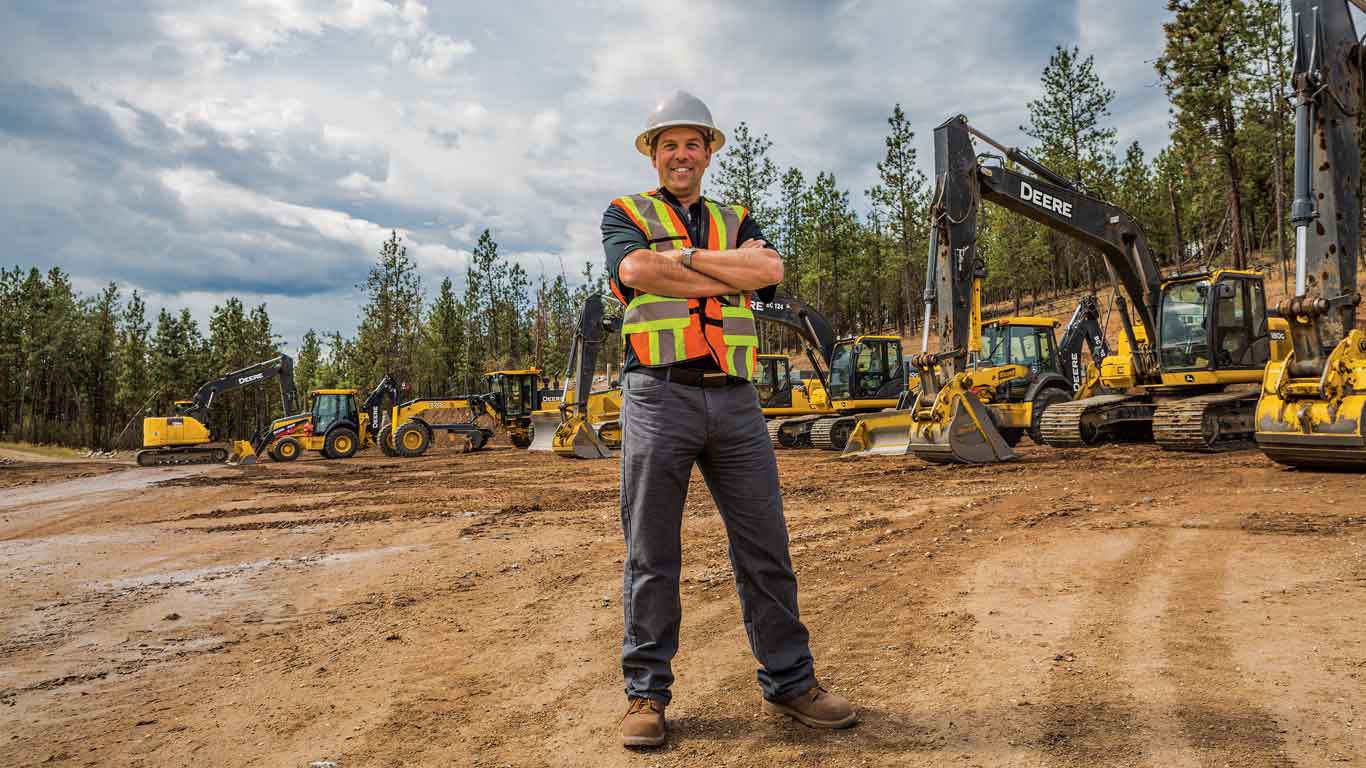 Man with construction hat and vest standing at a worksite with John Deere Construction equipment lined up behind him