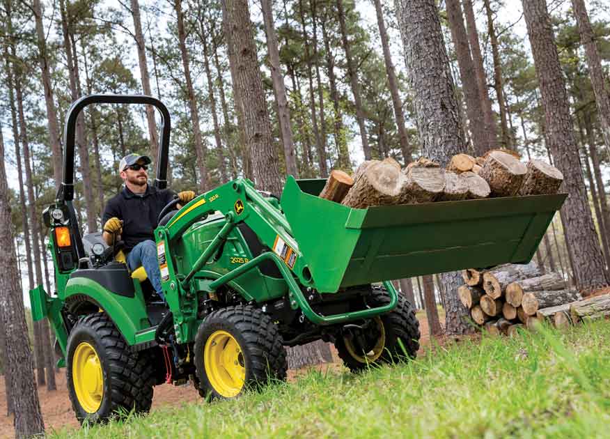 Man operating a John Deere 2025R Compact Tractor with bucket attachment carrying logs