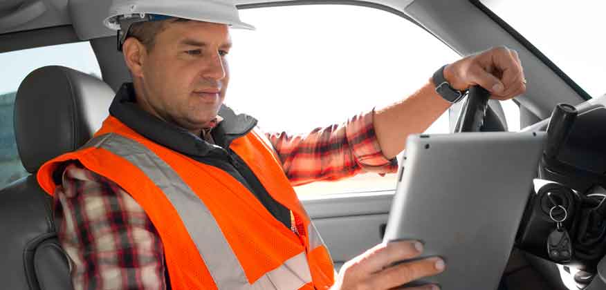 Man in orange construction vest and white hard hats sitting in a vehicle looking at an iPad