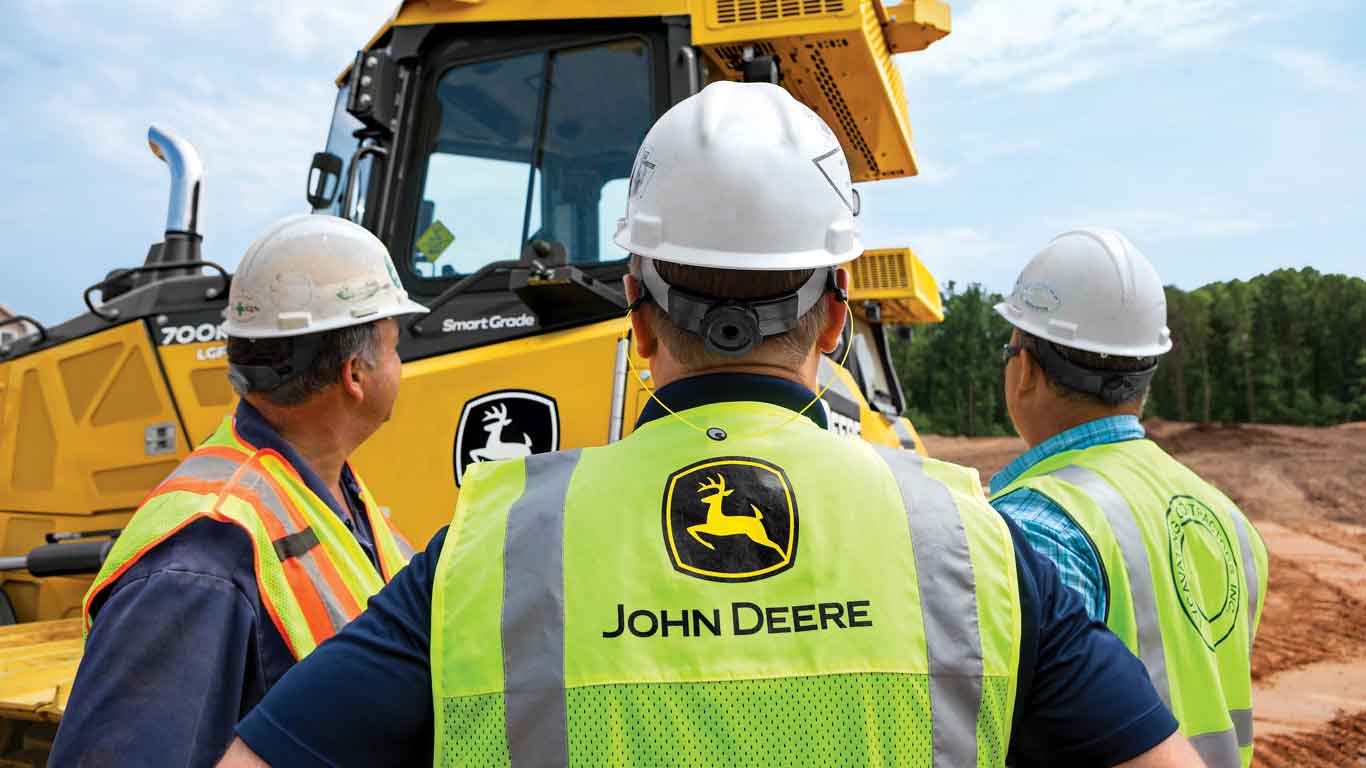 Three men with hard hats standing at a worksite with John Deere Construction equipment in the background