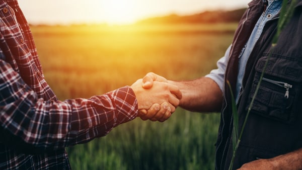 Two people shake hands in a field at sunset, one in plaid, the other in a vest.