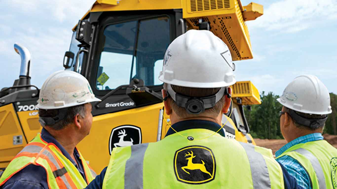 Three men in construction gear near John Deere construction equipment at a worksite