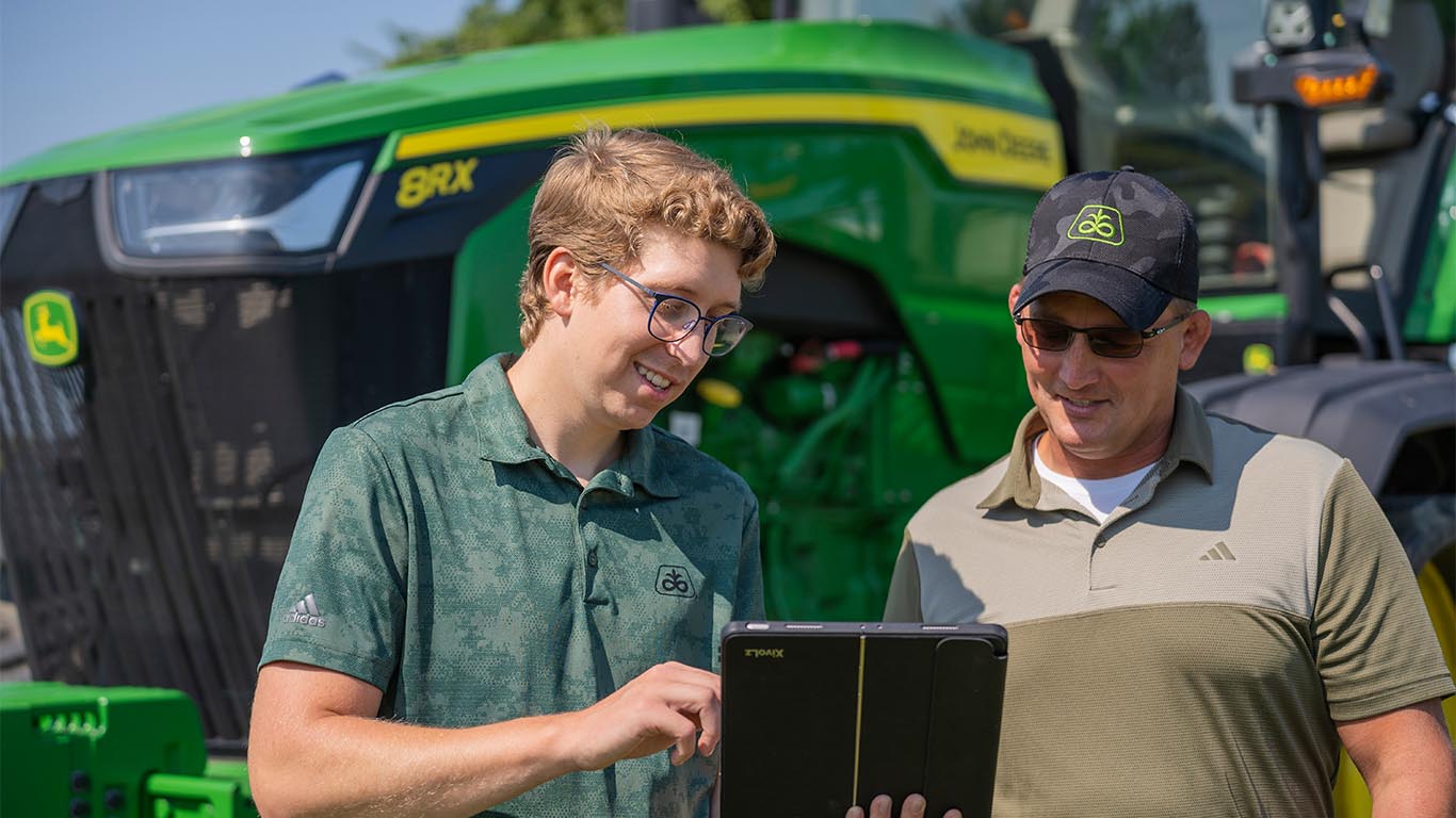 two farmers viewing a tablet in front of an 8RX Deere tractor