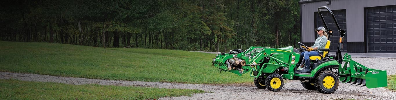 Woman driving a 1025R Compact Tractor with logs in its front grapple.