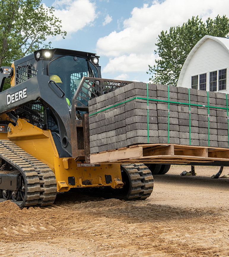 325G Compact Track Loader with forks attachment hauls a load of landscaping blocks in front of a white barn.