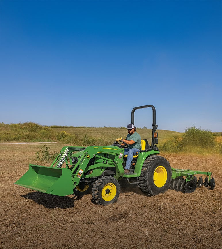 A woman driving a John Deere 3E Series Tractor on a rural property.