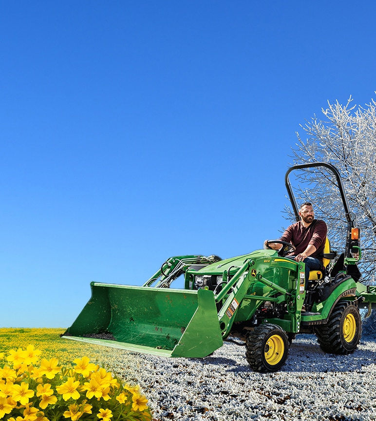 A man driving a John Deere 1025R Compact Tractor with loader attachment from snow-covered grass to blooming fields.