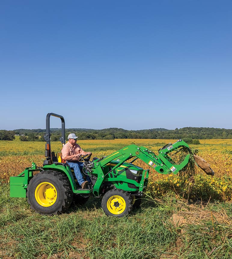 A man using a John Deere compact utility tractor  with a grapple attachment to clear up a pasture.