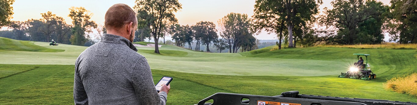 a golf superintendent looks at his phone while golf crew mows the golfing greens as the sun rises.