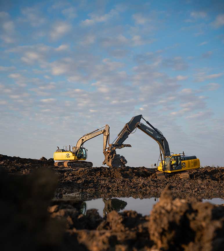 Two large yellow John Deere excavators digging a trench in a brown field