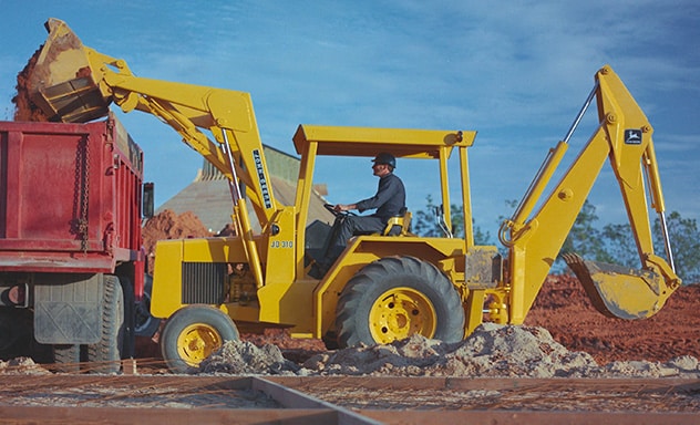 A person operating a John Deere JD 310 Backhoe dumping dirt into a red truck at a work site.