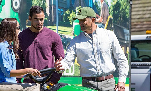 A dealer talking to a couple as a woman sits on a John Deere riding mower.  