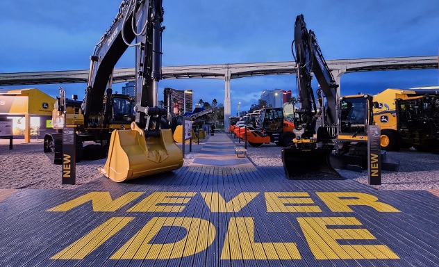 Two John Deere excavators shown with "NEVER IDLE" campaign slogan painted on ground in front of the buckets