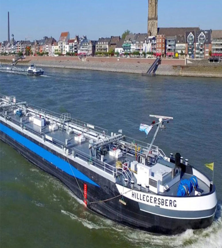 A long blue black and white boat drives across the blue water. With buildings, houses and another boat in the background.