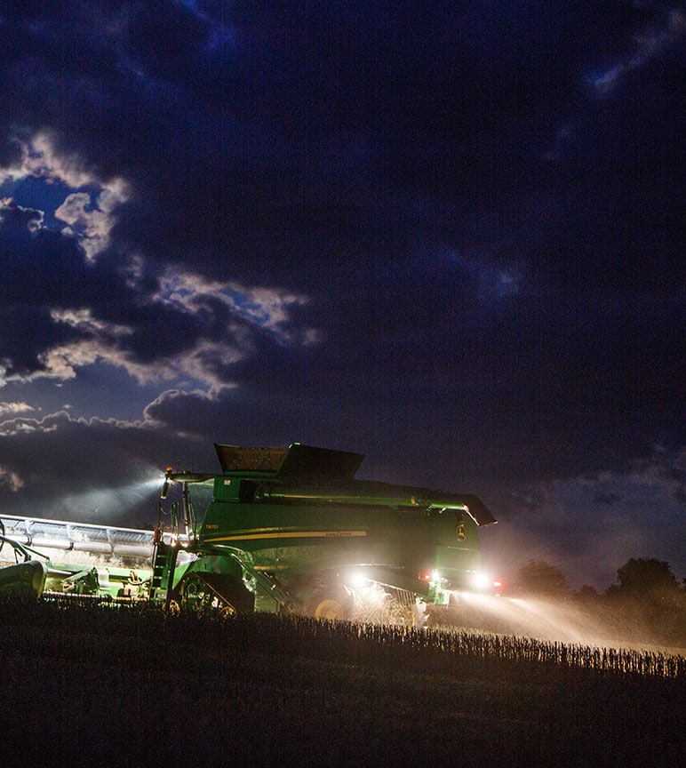 John Deere combine's lights shining as it harvests a dark field