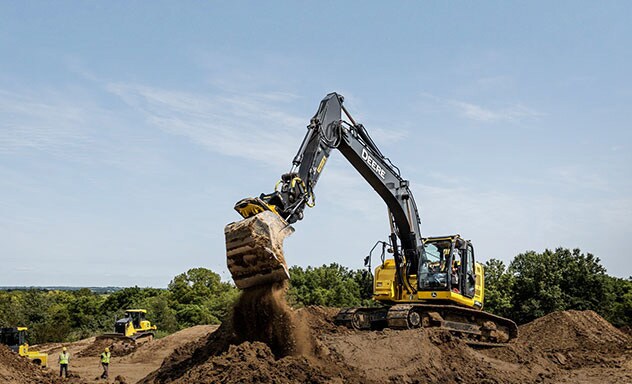 John Deere excavator sits on a dirt bench and dumps a shovel of earth.