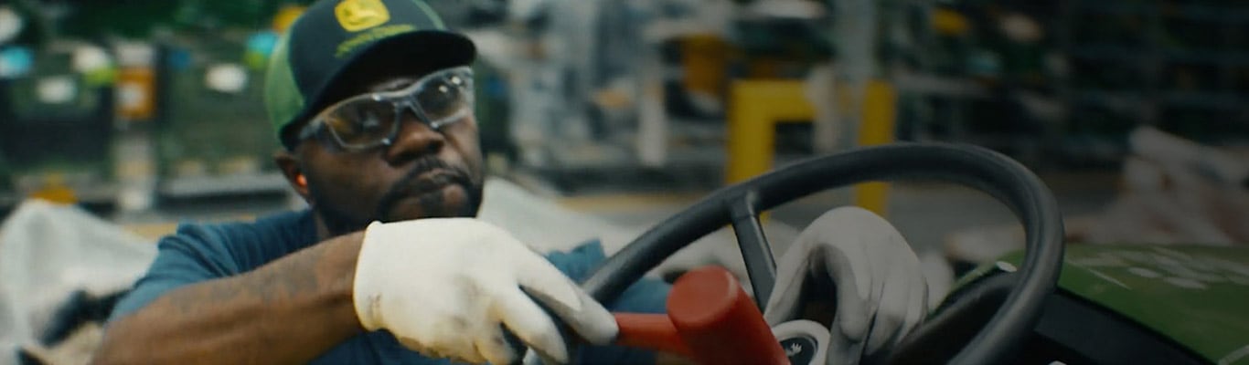 Man in a factory setting; he is hammering a green piece of John Deere equipment.