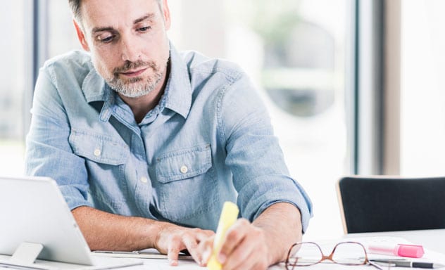 Man highlighting financial reports with laptop on desk nearby.