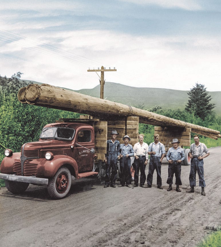 Vintage photo of six loggers in the stand in front of a pick-up truck with three felled trees strapped to the top.