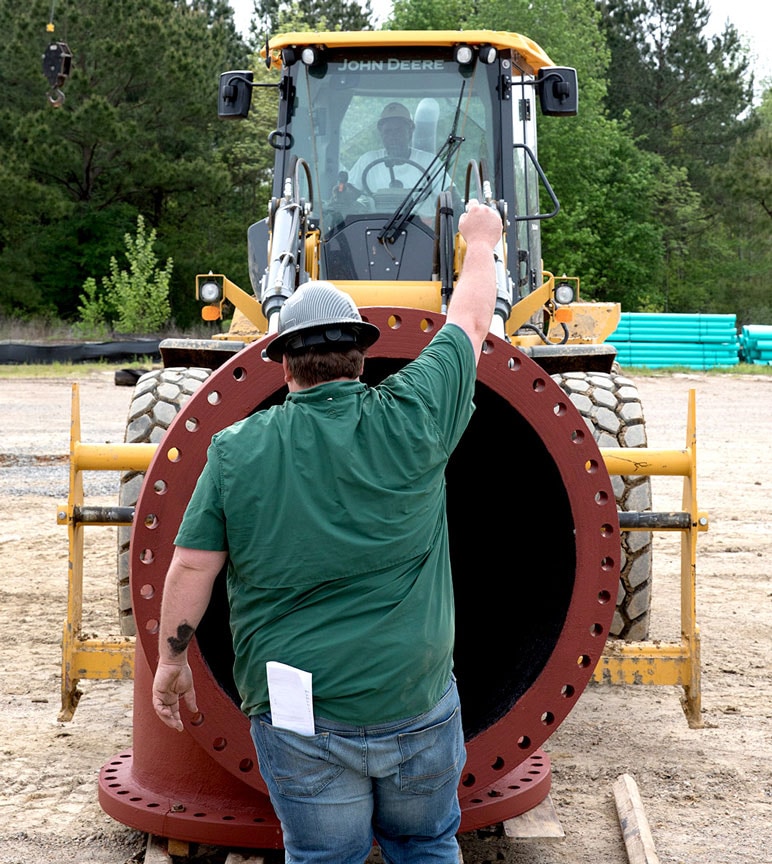 A wheel loader operator moves a section of pipe while another worker directs the placement. 