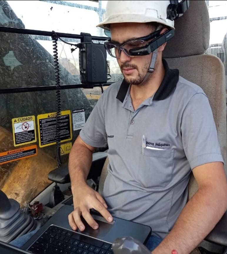 A man wearing a white construction hat is in operating a computing with smart glasses on, inside the cab of a machine.