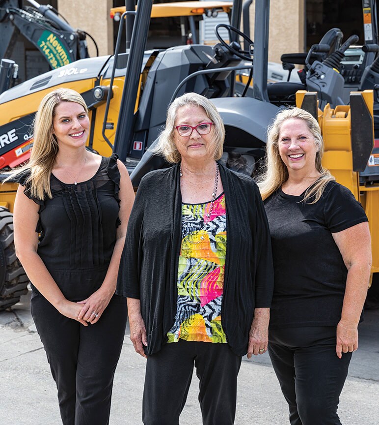 Amanda Matson, Janet Ferguson, and Nikki Worden of Newman Backhoe Service stand in front of a John Deere 310L Backhoe.