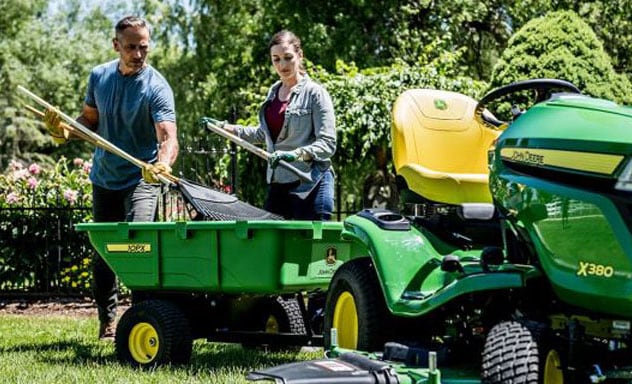 Man and woman holding rakes loading yard debris into a cart attached to a John Deere X380 riding lawn tractor.