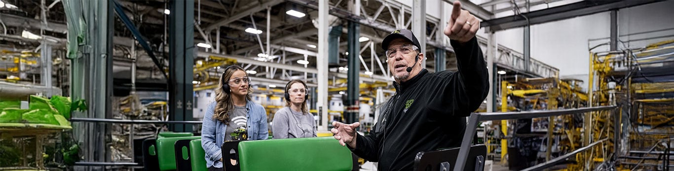 A man gestures while explaining something to two women, seated on green benches in a factory environment.