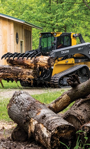 a John&nbsp;Deere 331 P-Tier CTL equipped with an RR84 Root Rake to move logs by a garage