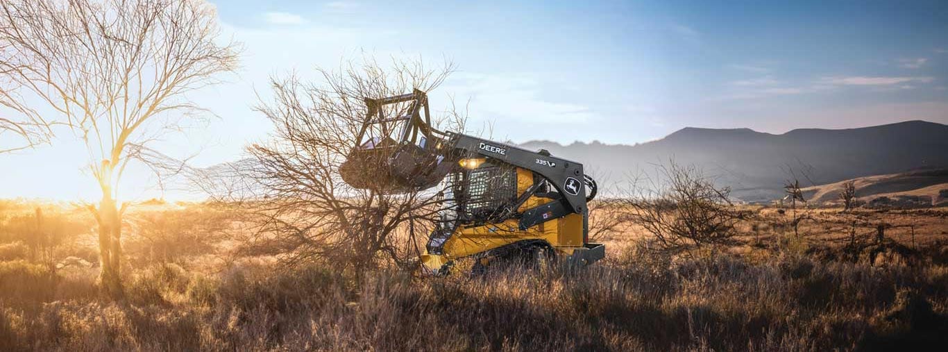 John Deere compact track loader clears brush in dry field at sunset.