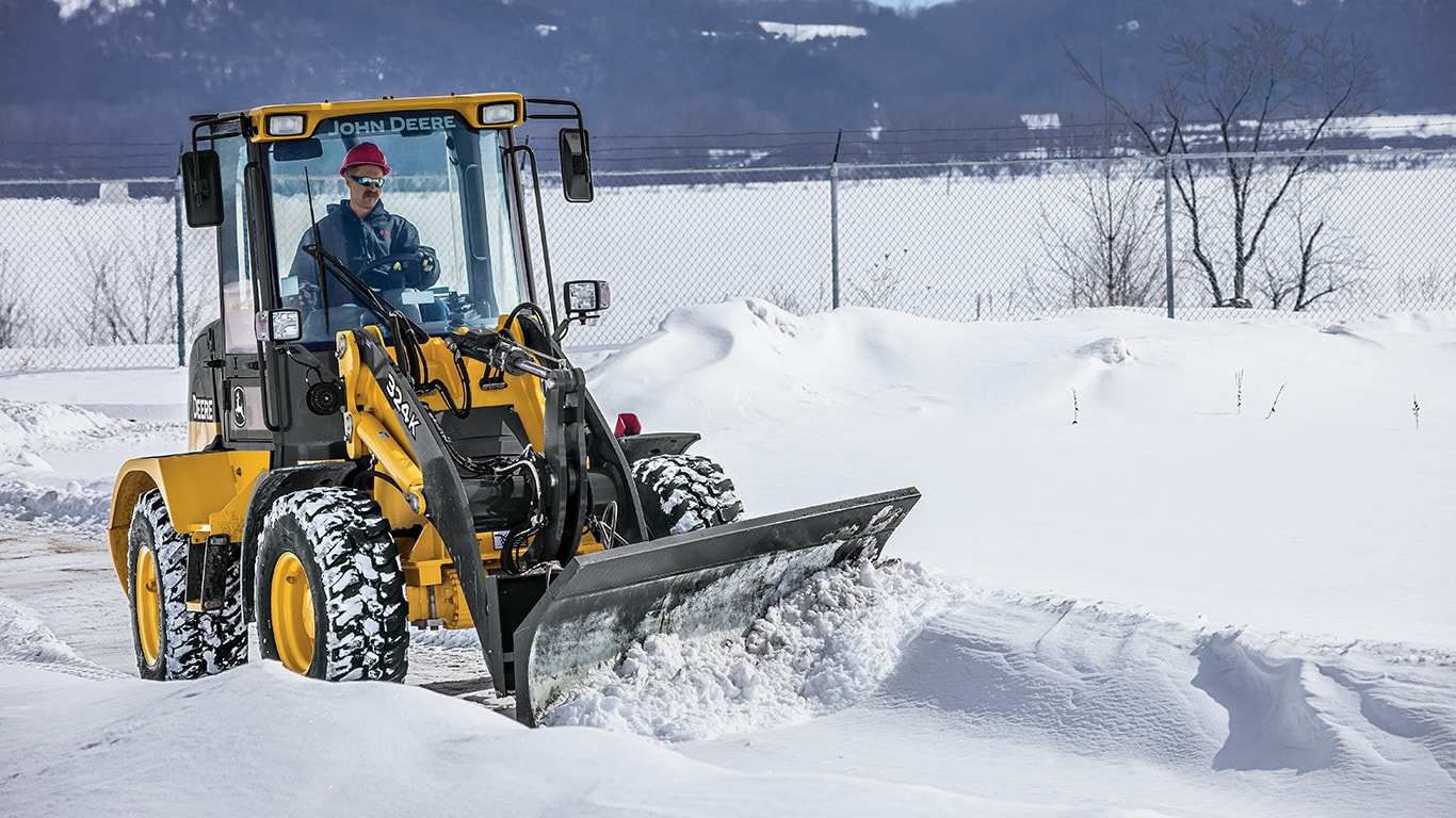 Cargador Compacto 324K con cuchilla para nieve empujando la nieve de la carretera.