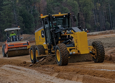 : The word “roadbuilding” is on top of a John Deere SmartGrade Motor Grader and Wirtgen HAMM tandem roller working together at a roadbuilding site. 