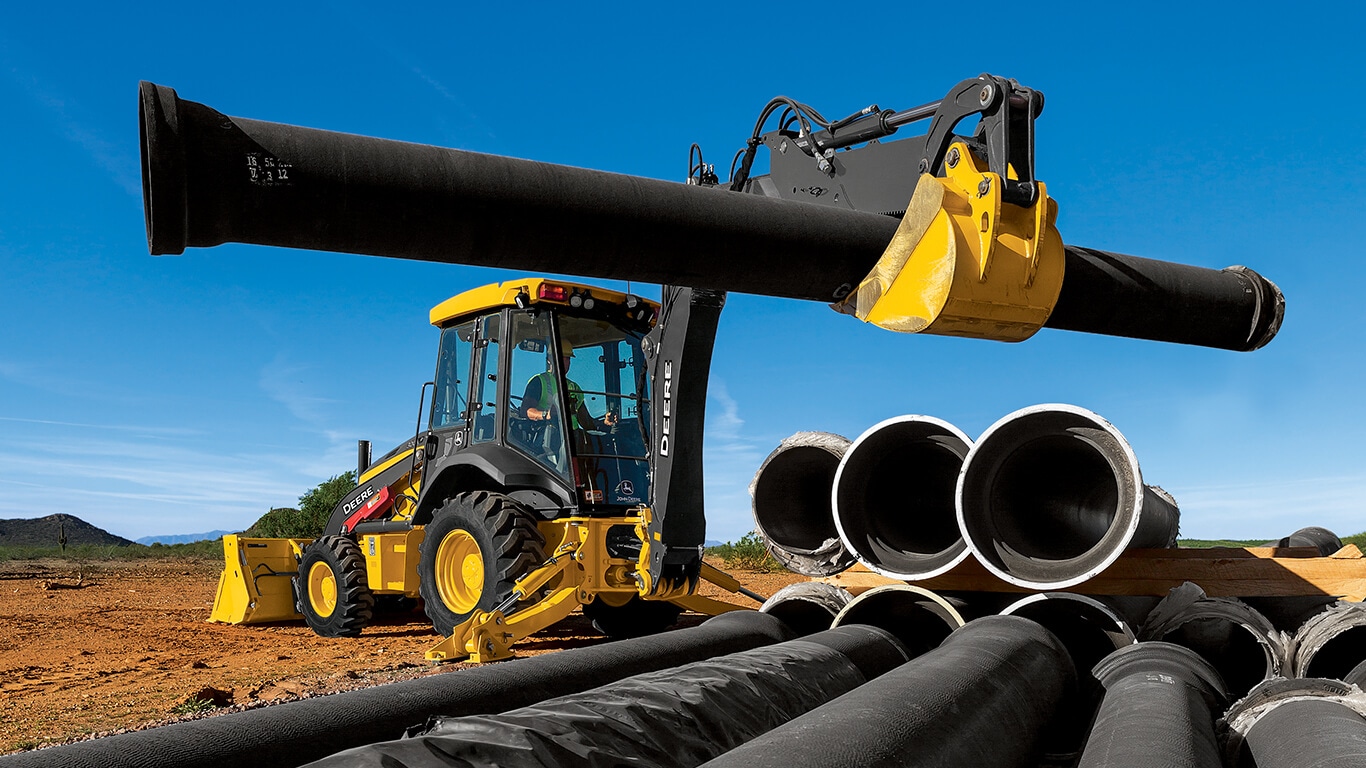 A backhoe using the bucket to lift a large pipe on a job site.