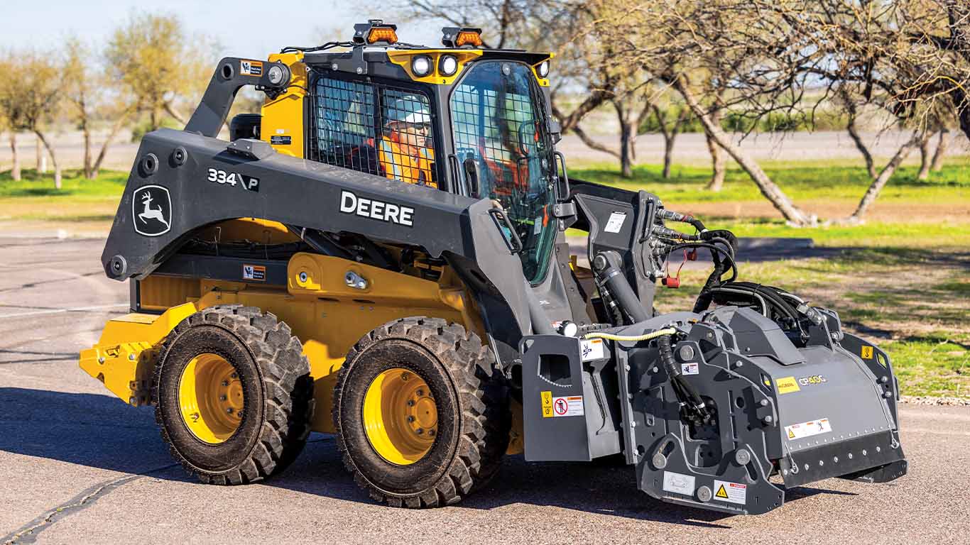 A John Deere 334 P-Tier skid steer loader with a cold planer attachment is in motion at a construction site.