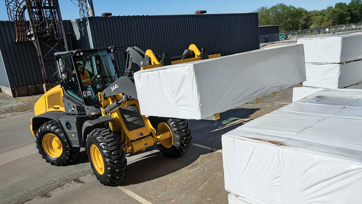 A John Deere 344L Compact Wheel Loader shown working in a tight space to load pallets.