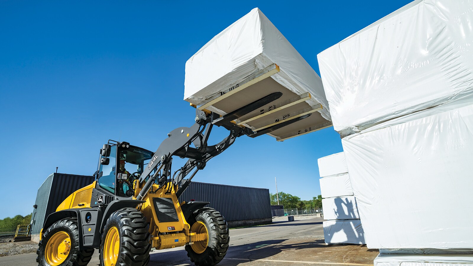 344L High Lift Wheel Loader lifting a pallet of wrapped goods up onto a stack well above the loader’s cab