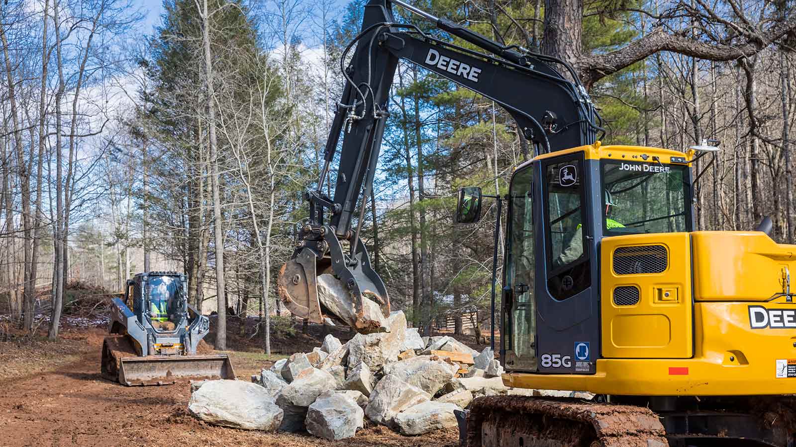 A John Deere 85G Mid-Size Excavator moves rocks with the assistance of a John Deere 333G Compact Track Loader