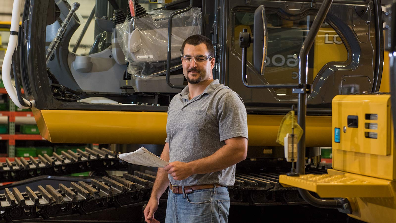 Jacob Henderson, a Product Development Process Design Engineer, shown working in the Deere-Hitachi factory in front of a John Deere excavator